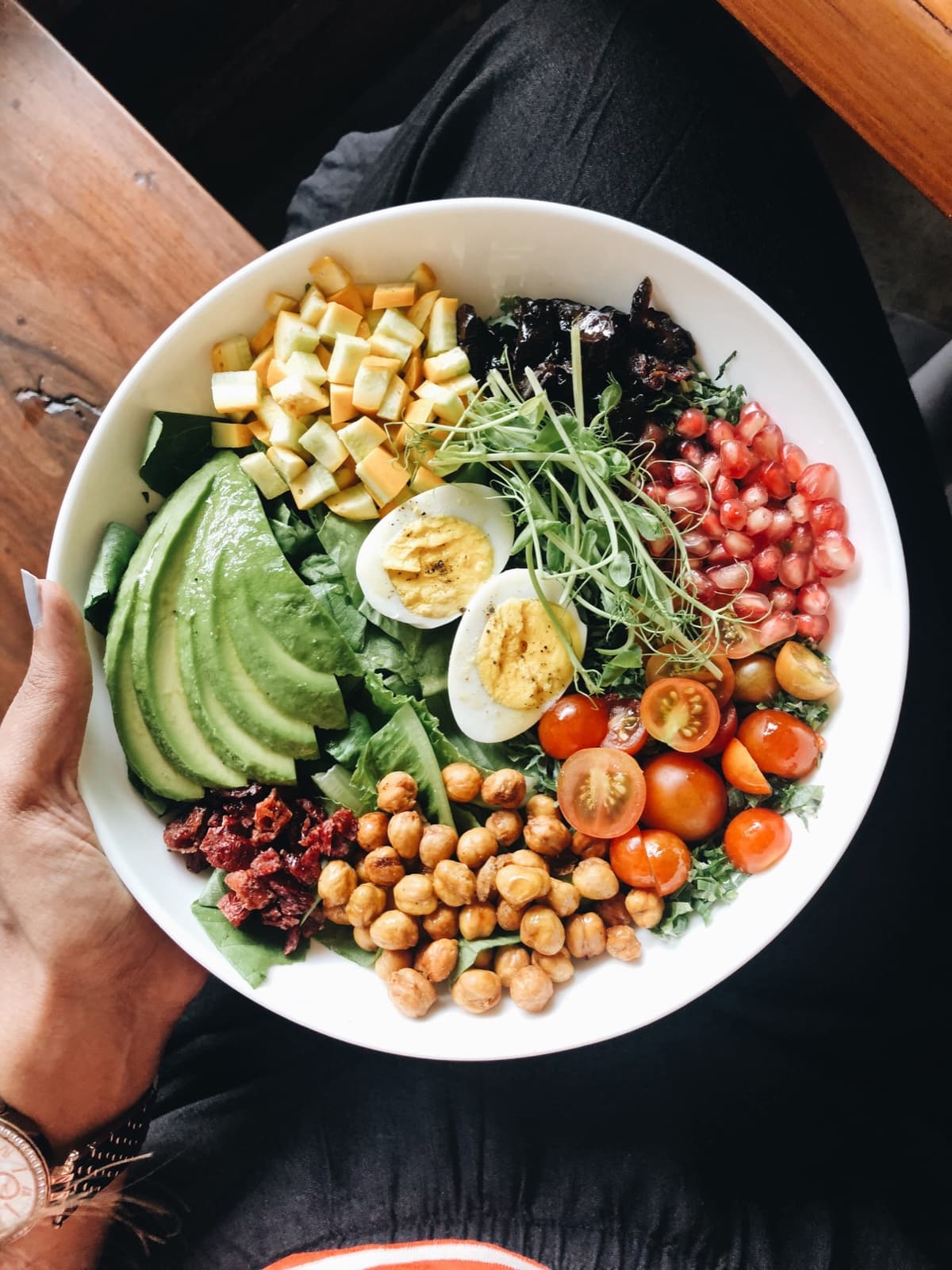 A bowl of greens, eggs, avocado, chickpeas and tomatoes held over a wooden table